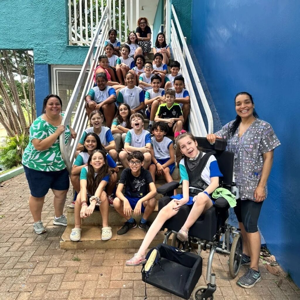 A imagem mostra um grupo de cerca de 20 crianças e 3 adultas reunidas em uma escadaria externa. A maioria das crianças veste uniformes escolares compostos por camisetas brancas com mangas azuis e verdes. No centro da imagem, em primeiro plano, há uma criança em uma cadeira de rodas adaptada, usando uma camiseta branca com detalhes azuis e um colar cervical cinza. As duas mulheres adultas estão posicionadas nas laterais da escadaria: uma à esquerda, usando uma blusa verde estampada e shorts azuis, e outra à direita, vestindo um jaleco cinza estampado com leggings pretas. A escadaria tem corrimãos brancos e está situada entre uma parede azul vibrante à direita e uma parede verde texturizada à esquerda. O chão é pavimentado com tijolos intertravados de cor avermelhada. A iluminação é natural e intensa, indicando que a foto foi tirada durante o dia. Todos parecem estar felizes, muitos sorrindo para a câmera, transmitindo um clima de alegria e inclusão. Algumas camisetas exibem a palavra "Piracicaba". A imagem sugere um ambiente escolar ou de atividade comunitária.