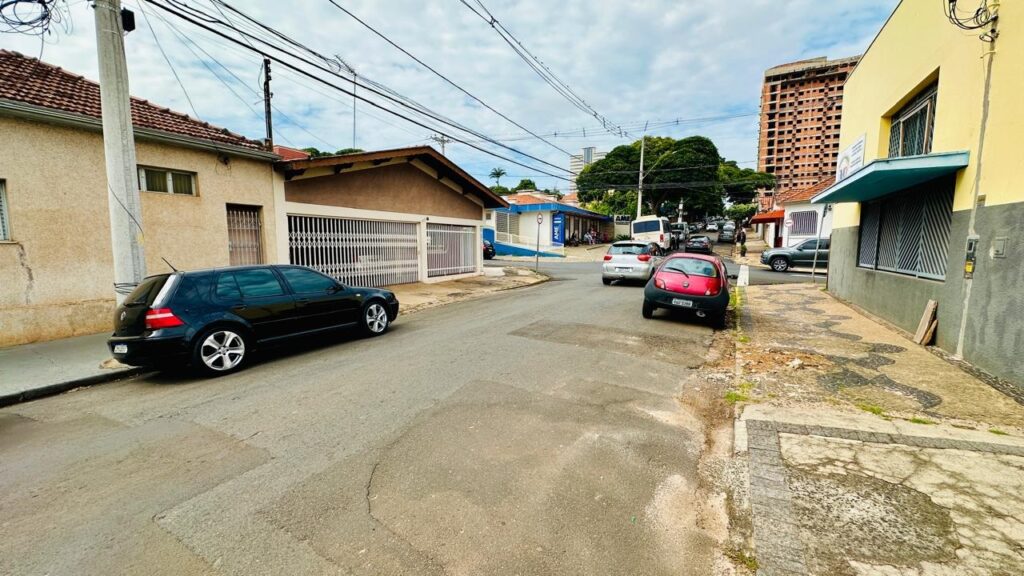 A imagem mostra uma cena urbana em uma rua pavimentada, provavelmente em um bairro residencial ou misto com comércios. Ao lado esquerdo da imagem, há um carro preto estacionado — um Volkswagen Golf — e do lado direito, mais à frente, há um carro vermelho, que parece ser um Ford Ka, também estacionado. A rua tem uma leve inclinação e está ladeada por calçadas. Ao fundo, é possível ver algumas árvores verdes e uma construção alta de tijolos aparentes que parece estar em andamento. Também há um prédio azul com a palavra "AME" escrita na fachada, sugerindo que pode ser algum tipo de estabelecimento comercial ou serviço. No céu, há muitos fios de energia elétrica cruzando a imagem, o que é comum em áreas urbanas. A iluminação indica que o dia está nublado ou com luz difusa, sem sombras muito fortes. No geral, a imagem transmite uma sensação de um bairro tranquilo, com trânsito leve e uma mistura de construções residenciais e comerciais.