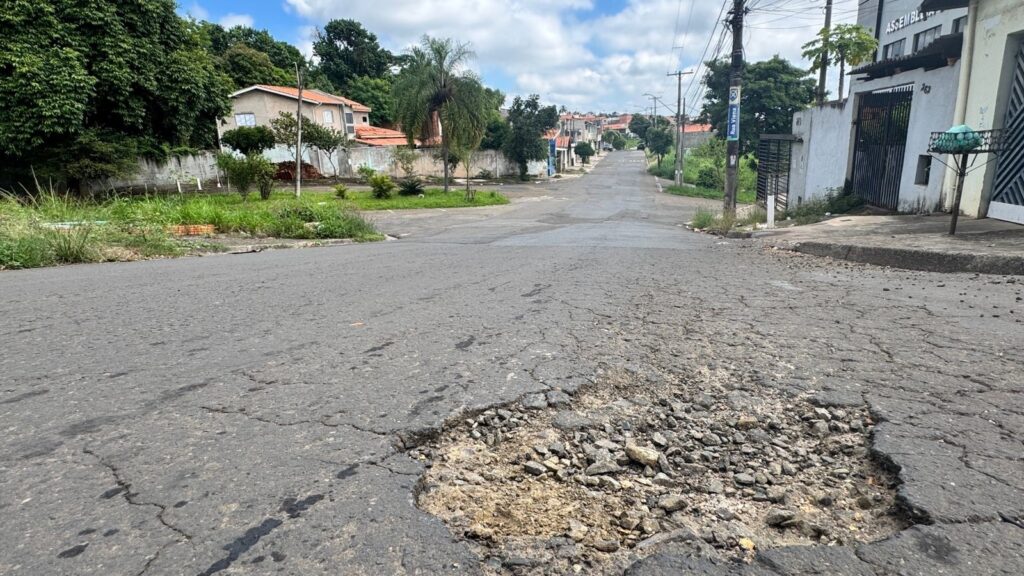 A imagem mostra uma rua residencial com um grande buraco no asfalto em primeiro plano, ocupando boa parte da visão próxima. O buraco é profundo e irregular, com pedras soltas e terra visível dentro dele, indicando desgaste e falta de manutenção. A rua é ladeada por casas simples, com telhados de cerâmica vermelha, e postes de energia elétrica com fios aparentes estão ao lado direito da via. No poste próximo ao buraco, há uma placa azul com o nome da rua, "Rua Viena". Ao fundo, o céu está parcialmente nublado, com nuvens brancas sobre um céu azul claro, sugerindo um dia ensolarado. Também se vê uma lixeira metálica no lado direito da rua. A perspectiva da foto é baixa e próxima ao chão, destacando o buraco e dando uma sensação da profundidade do dano na via. A cena transmite a ideia de um bairro tranquilo, porém com problemas na infraestrutura da rua.