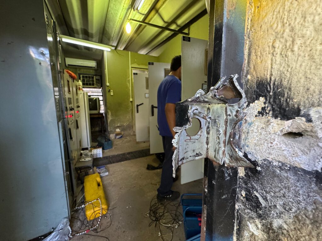 A imagem mostra uma sala industrial ou técnica, possivelmente uma subestação elétrica ou sala de controle, vista através de uma porta ou abertura com uma trava de metal danificada em primeiro plano. A trava está rasgada e com bordas irregulares, indicando que foi forçada ou danificada. No interior da sala, há painéis elétricos com luzes vermelhas acesas e diversos equipamentos espalhados pelo chão, incluindo uma caixa amarela e uma caixa azul. Um homem, vestido com camiseta azul e calça escura, está de costas, olhando para uma porta ou painel branco ao fundo da sala. O ambiente é iluminado por luz artificial, com tons amarelados e verdes, e as paredes são de cor verde escura, com teto metálico ondulado. O chão é de concreto, e o espaço transmite uma sensação de desordem e possível emergência ou reparo em andamento.