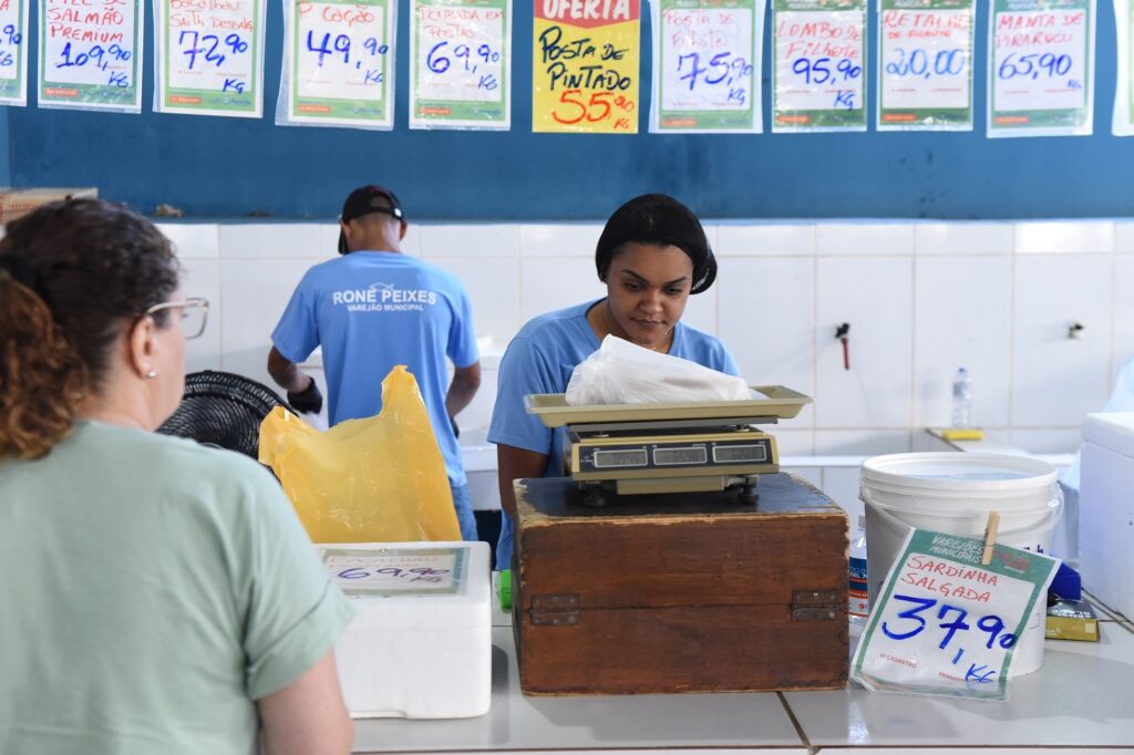 A imagem mostra uma cena dentro de um mercado municipal de peixes. No centro, uma mulher vestindo uma camiseta azul está pesando um saco plástico branco em uma balança digital sobre um balcão branco. Ao lado dela, um homem também de camiseta azul está de costas, aparentemente trabalhando no mesmo local. Na frente, um cliente, com cabelo cacheado e óculos, observa a pesagem. Ao fundo, uma parede azul exibe várias placas com os nomes e preços de diferentes tipos de peixes, como salmão, pintado, filhote e bacalhau, com valores em reais por quilograma. Ao lado direito do balcão, há um balde branco com uma placa indicando o preço da sardinha salgada. O ambiente é típico de um varejão municipal, com uma atmosfera simples e funcional.