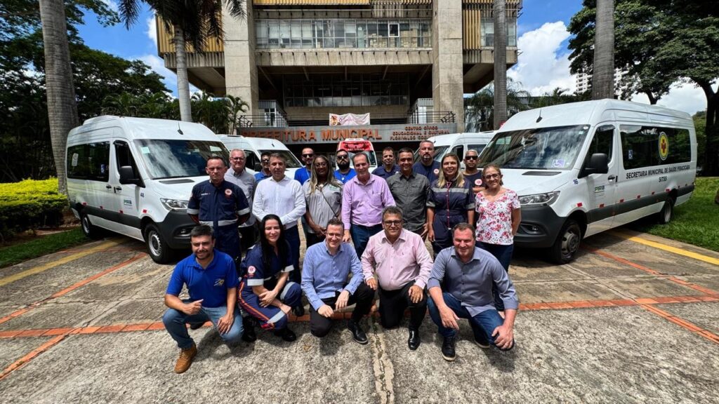 A imagem mostra um grupo de cerca de 18 pessoas posando para uma foto em frente a um edifício público identificado como "Prefeitura Municipal" e "Centro Cívico Educacional e Cultural Florivaldo Coelho". O grupo está distribuído em três fileiras, com algumas pessoas agachadas na frente e outras em pé atrás. Entre os presentes, há profissionais vestidos com uniformes médicos azul escuro com detalhes em vermelho e branco, além de outras pessoas com roupas casuais, como camisas sociais e blusas. Ao redor do grupo estão estacionadas três vans brancas da marca Mercedes-Benz Sprinter, sendo que duas delas estão mais próximas e visíveis nas laterais da foto. Uma das vans à direita traz a inscrição "Secretaria Municipal de Saúde Piracicaba" e exibe um símbolo de cruz vermelha, indicando que são veículos destinados à área da saúde. O cenário é externo, com um céu claro e parcialmente nublado, árvores e palmeiras ao fundo, e a iluminação natural do dia realça a cena, criando sombras no chão. A atmosfera geral é profissional e comemorativa, sugerindo que a foto foi tirada para marcar a entrega ou apresentação desses novos veículos de transporte médico à comunidade.