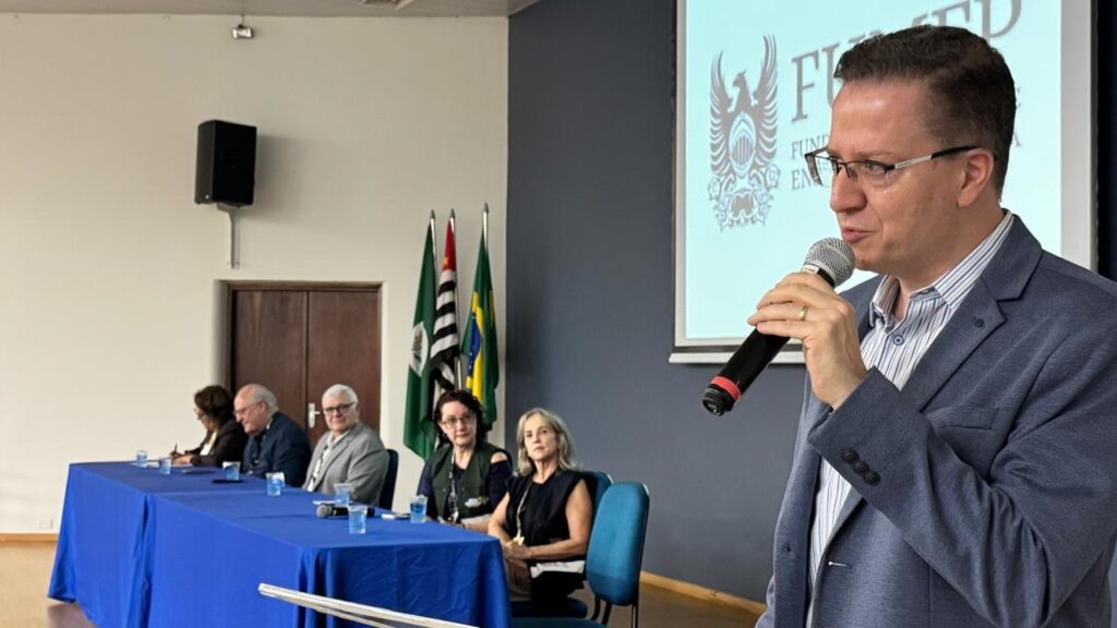 A imagem mostra um evento formal em um ambiente interno, possivelmente uma conferência ou palestra. Em primeiro plano, à direita, há um homem de meia-idade vestindo um blazer azul e camisa listrada, falando ao microfone. Ele parece ser o palestrante ou apresentador principal. Ao fundo, há uma mesa longa coberta com um pano azul, onde cinco pessoas estão sentadas, ouvindo atentamente. Entre elas, há homens e mulheres de diferentes idades. Atrás da mesa, estão posicionadas três bandeiras, sendo uma delas a bandeira do Brasil e outra a bandeira do estado de São Paulo, sugerindo que o evento ocorre no Brasil. Na parede ao fundo, há uma tela de projeção que exibe um logo com as letras "F" e "FUND", possivelmente relacionado ao tema ou à organização do evento. O ambiente é bem iluminado, com paredes brancas e detalhes em azul escuro. Um alto-falante está fixado na parede à esquerda, indicando que o local é preparado para eventos com áudio. No centro da mesa, há copos plásticos transparentes com água, indicando que o evento pode durar algum tempo.