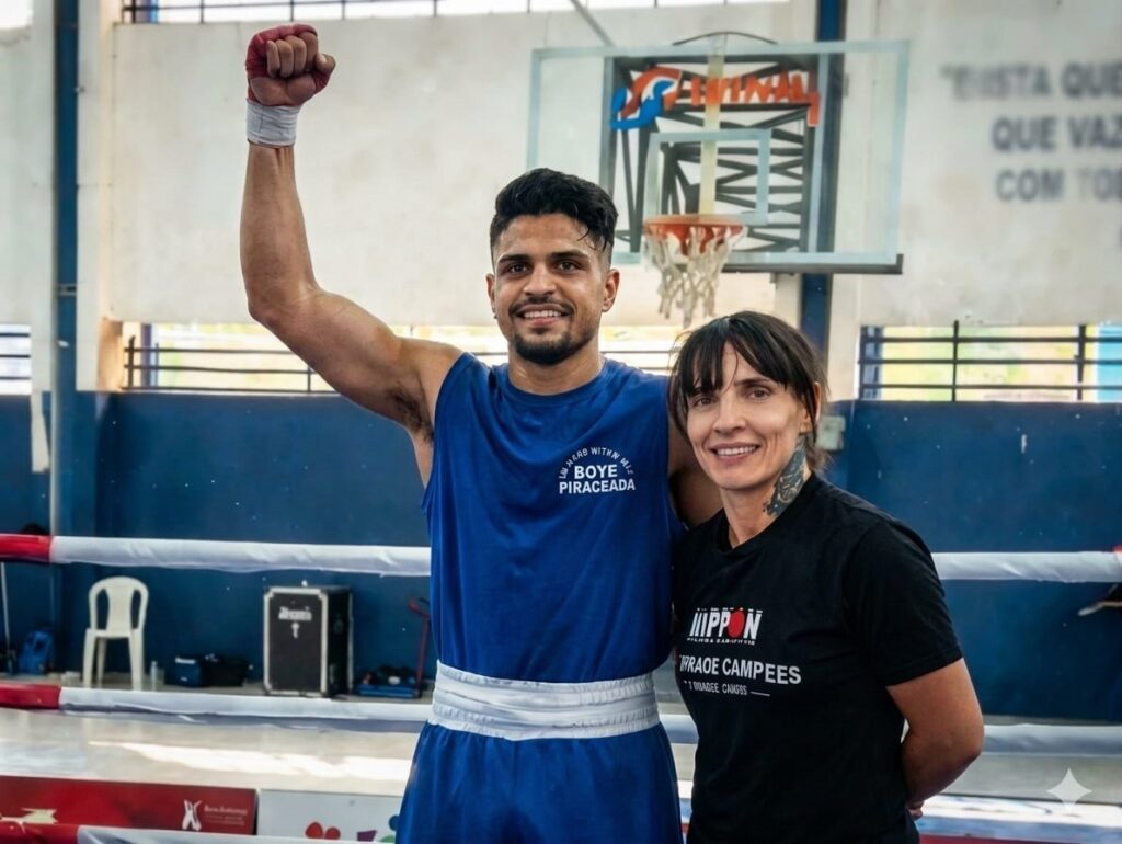 A imagem mostra um momento de celebração em um ambiente esportivo fechado, provavelmente uma academia de boxe ou centro esportivo. No centro da foto, há um jovem levantando o braço direito com o punho fechado, em sinal de vitória. Ele veste uma regata azul com a inscrição "BOYE PIRACICABA" e shorts azuis com detalhes brancos. Suas mãos estão envoltas em bandagens e fitas vermelhas, típicas de boxeadores. Ao lado dele, uma mulher sorri, vestindo uma camiseta preta com as palavras "IPPON" e "PRAÇA DE CAMPEÕES". Ela tem um tatuagem visível no pescoço. Ao fundo, é possível ver um ringue de boxe com cordas vermelhas e brancas, uma cadeira plástica branca e uma parede com janelas e algumas inscrições em português. Também há uma cesta de basquete fixada na parede. A cena transmite uma sensação de vitória e comemoração, com iluminação natural que destaca os personagens principais.