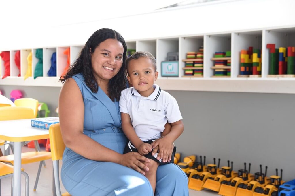 A imagem mostra uma mulher e um menino sentados juntos em um ambiente que parece ser uma sala de aula ou um espaço de recreação infantil. A mulher tem cabelos longos, cacheados e escuros, está sorrindo de forma acolhedora e veste um macacão azul sem mangas. Ela está sentada e segura o menino no colo. O menino, que aparenta ter cerca de 3 ou 4 anos, usa uma camisa polo branca com detalhes pretos na gola e shorts escuros. Ele olha diretamente para a câmera com uma expressão neutra. Ao fundo, há uma estante branca com vários compartimentos, onde estão organizados brinquedos coloridos, como blocos de montar e livros. Abaixo da estante, no chão, há uma fileira de carrinhos de brinquedo amarelos. À esquerda, é possível ver uma mesa amarela com algumas cadeiras ao redor e uma caixa azul sobre a mesa. A iluminação é clara e suave, criando um ambiente alegre e convidativo, típico de uma creche ou escola infantil.