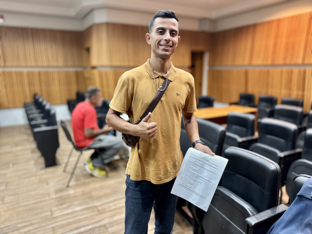 A imagem mostra um homem sorridente, vestindo uma camisa polo amarela e jeans, segurando um currículo impresso em uma das mãos e fazendo um gesto de positivo (joinha) com a outra. Ele está em uma sala de auditório com cadeiras pretas e paredes de madeira clara. Ao fundo, outra pessoa está sentada, vestindo camiseta vermelha e calça cinza, olhando para frente. O homem parece estar feliz e confiante, possivelmente por ter conseguido uma oportunidade ou estar participando de um evento relacionado a emprego ou educação. O currículo que ele segura está escrito em português e destaca sua formação acadêmica, cursos complementares e experiências profissionais na área administrativa e logística.