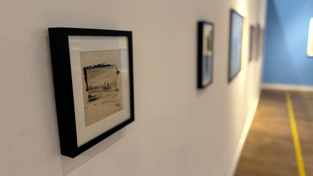 A imagem mostra uma galeria de arte com várias obras emolduradas penduradas na parede branca. A foto foi tirada de um ângulo lateral, fazendo com que as molduras apareçam em perspectiva, com a primeira obra em foco nítido e as demais se tornando progressivamente desfocadas. A obra mais próxima parece ser um desenho ou esboço com um tema náutico, possivelmente um barco à vela sobre a água. Ao fundo, há uma parede azul que adiciona um toque de cor ao ambiente predominantemente neutro. No chão, uma linha amarela delimita a área para observação das obras. A iluminação é suave e direcionada, destacando as obras expostas e criando um ambiente tranquilo e elegante. A disposição das molduras é uniforme, sugerindo uma exposição organizada e profissional.