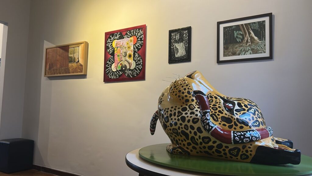 A imagem mostra um ambiente de galeria de arte com várias obras expostas em uma parede de tom claro, provavelmente branca ou bege. Em destaque, na frente, há uma escultura grande e colorida de um jaguar estilizado, feita possivelmente de cerâmica ou resina. O jaguar é amarelo com manchas pretas características, e uma cobra vermelha com detalhes brancos está enrolada em seu corpo. A escultura está sobre um pedestal circular verde, e o jaguar parece estar olhando para cima. Ao fundo, na parede, estão penduradas quatro obras emolduradas. Da esquerda para a direita: uma pintura horizontal emoldurada em madeira mostrando uma paisagem, possivelmente vista através de uma janela; uma obra quadrada com uma moldura vermelha contendo elementos abstratos coloridos e padrões circulares em preto e branco; uma pequena obra vertical em preto e branco; e uma pintura de paisagem que mostra uma floresta com troncos de árvores grandes. A iluminação é quente e direcionada, típica de galerias, criando sombras suaves e destacando as texturas das obras. A perspectiva da foto é baixa, valorizando a escultura em primeiro plano, enquanto as pinturas ao fundo complementam o cenário. A escultura tem uma aparência lisa e brilhante, contrastando com a parede de acabamento fosco.