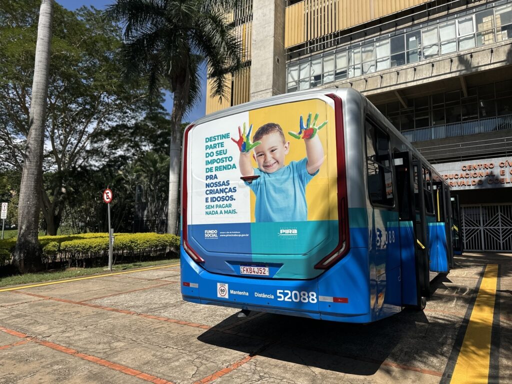 A imagem mostra a parte traseira e lateral de um ônibus azul e prata estacionado em frente a um prédio. Na lateral do ônibus há um grande anúncio publicitário com a foto de uma criança sorridente, que segura as mãos abertas, com as palmas pintadas de várias cores. O texto do anúncio, em português, incentiva as pessoas a destinarem parte do seu imposto de renda para ajudar crianças e idosos, ressaltando que isso não gera custo adicional ("SEM PAGAR NADA A MAIS"). No anúncio também aparecem os logotipos do Fundo Social e da Prefeitura de Piracicaba, além do endereço do site "piracicaba.sp.gov.br". O ônibus tem o número 52088 e a placa FKB4J52. Ao fundo, vê-se um prédio público com a inscrição "CENTRO CÍVICO EDUCACIONAL E CULTURAL RIVALDO COELHO" (parte do texto está parcialmente coberta). O ambiente parece ser uma área urbana com árvores e bastante luz natural, indicando um dia ensolarado.