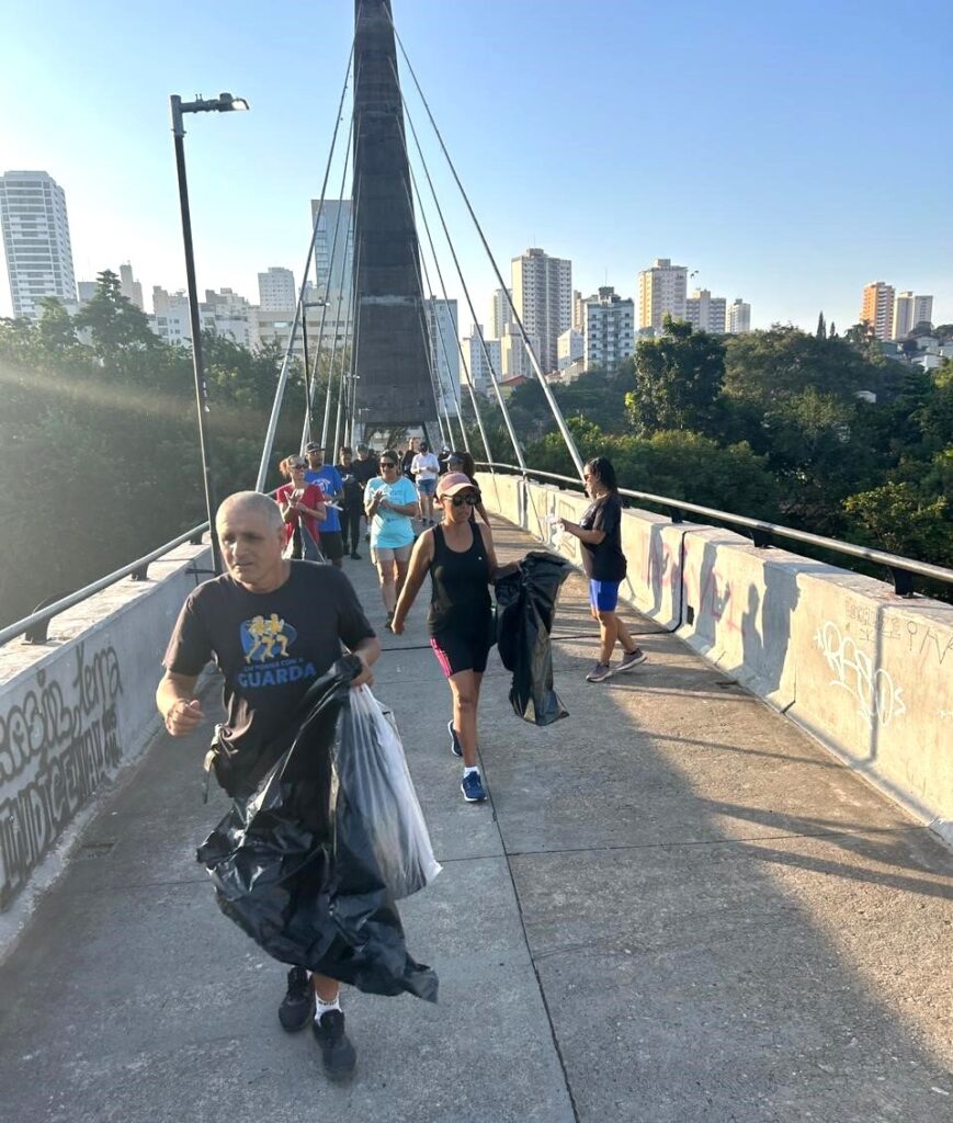 A imagem mostra um grupo de pessoas caminhando sobre uma ponte, aparentemente participando de uma atividade de limpeza ambiental. No primeiro plano, um homem carrega sacos pretos de lixo, enquanto outras pessoas ao fundo também seguram sacos semelhantes. A ponte é uma estrutura moderna, com cabos de sustentação e um pilar central alto, que domina a composição da foto. A luz do sol da manhã ilumina a cena, criando sombras longas e ressaltando os detalhes da ponte e das roupas das pessoas. As pessoas estão vestidas com roupas casuais e esportivas, e parecem concentradas na tarefa de recolher o lixo. O ambiente ao redor mostra algumas árvores e edifícios ao longe, sugerindo que a ponte está localizada em uma área urbana ou semiurbana. Na lateral da ponte, há grafites, que acrescentam um toque urbano à cena. No geral, a imagem transmite uma sensação de engajamento comunitário e responsabilidade ambiental, com pessoas unidas para limpar e cuidar do espaço público.