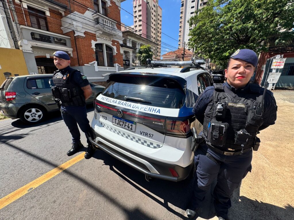 A imagem mostra dois policiais em pé ao lado de uma viatura policial branca. A viatura possui a inscrição "PATRULHA MARIA DA PENHA" na parte traseira, indicando que é uma unidade especializada no atendimento de casos relacionados à Lei Maria da Penha, que protege mulheres contra violência doméstica no Brasil. Os policiais estão vestidos com uniformes azul escuro, coletes táticos e boinas azuis. O policial à esquerda é um homem com bigode, e a policial à direita é uma mulher. Ambos estão em posição de atenção, com as mãos atrás das costas, olhando para a câmera. A viatura tem a placa "TJC7F41" e outros detalhes visíveis, como "VTR 57". Ao fundo, é possível ver uma rua urbana com algumas árvores, um prédio de tijolos vermelhos e edifícios mais altos, indicando que a cena ocorre em uma área urbana. A iluminação é forte, sugerindo que a foto foi tirada durante o dia, provavelmente ao meio-dia. No geral, a imagem transmite uma sensação de segurança e vigilância, mostrando a presença policial especializada no combate à violência doméstica.