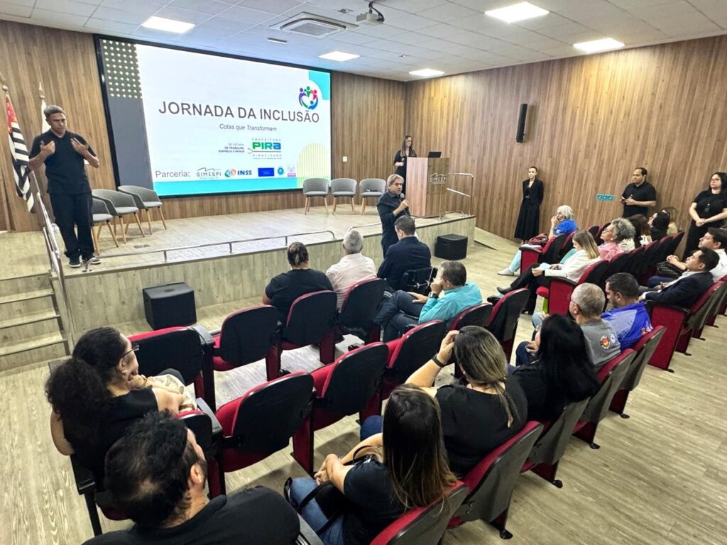 A imagem mostra um evento realizado em um auditório, provavelmente uma palestra ou conferência sobre inclusão, como indica o título no telão: "Jornada da Inclusão - Cotas que Transformam". No palco, um homem está falando para o público, usando uma camisa preta e gesticulando com as mãos. O telão atrás dele exibe o nome do evento, o logo da Prefeitura de Piracicaba, além de mencionar uma parceria com o SIMESPI e o INSS. À esquerda do palco, há uma bandeira do Brasil. O público está sentado em cadeiras vermelhas, prestando atenção ao palestrante. O ambiente é moderno, com paredes de madeira e iluminação clara, criando uma atmosfera profissional e acolhedora para discussão sobre inclusão social.