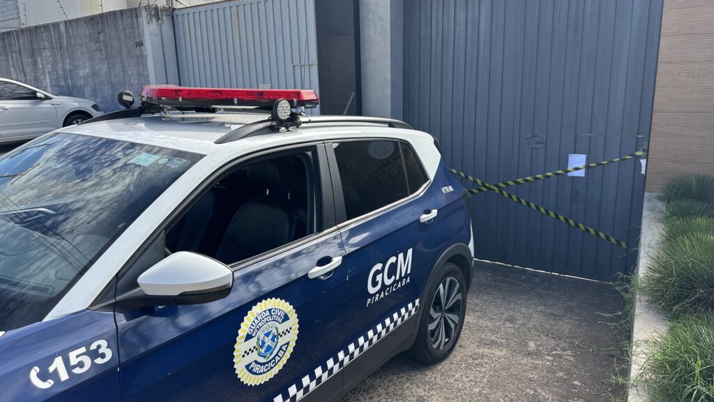 A imagem mostra uma viatura policial de cor azul com detalhes branco e amarelo, estacionada em frente um estabelecimento, que está interditado com fita amarela e preto.