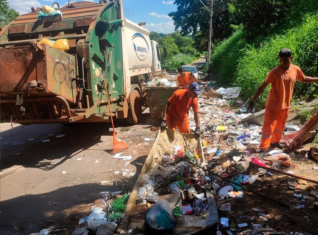 A imagem mostra uma cena de coleta e limpeza de lixo em um ambiente externo, possivelmente próximo a uma estrada ou área rural. No lado esquerdo da imagem, há um caminhão de lixo grande, predominantemente verde e branco, com a palavra "AMBIENTAL" visível na lateral. Perto do caminhão, há três trabalhadores da limpeza, todos vestidos com uniformes laranja e chapéus escuros, envolvidos na tarefa de recolher e organizar o lixo espalhado pelo chão. O chão está coberto por uma grande quantidade de resíduos variados, como sacos plásticos, garrafas e restos de comida. Um dos trabalhadores está agachado utilizando uma ferramenta para recolher o lixo, enquanto outro está em pé, também manipulando os resíduos. Ao fundo, há uma área com vegetação densa e verde, que contrasta com o lixo espalhado no solo. Na lateral do caminhão, também é possível ver um texto que diz "Reciclagem", seguido das frases "Por uma vida mais limpa" e "Por um planeta mais saudável", reforçando a mensagem de conscientização ambiental e incentivo à reciclagem. A cena transmite o esforço manual necessário para a limpeza e a importância da preservação ambiental.
