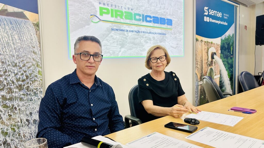 A imagem mostra duas pessoas sentadas lado a lado em uma mesa de reunião de madeira. À esquerda, um homem de cabelos grisalhos e óculos usa uma camisa azul estampada. À direita, uma mulher de cabelos curtos loiros e também de óculos, veste uma blusa preta com botões dourados nos ombros e segura uma caneta dourada em cima de documentos sobre a mesa. Ao fundo, há uma grande tela com o logo da "Prefeitura Piracicaba" e o texto "Secretaria de Habitação e Regularização Fundiária". À direita, um banner com o logo da "semae" e suas redes sociais está parcialmente visível. Nas extremidades da imagem, banners verticais mostram imagens relacionadas à água, como uma cachoeira e uma represa. Sobre a mesa, há diversos papéis, uma caneta roxa, um microfone, um celular preto e um chaveiro com controle remoto de carro. O ambiente parece ser uma sala de reuniões institucional, com iluminação clara e ambiente formal, sugerindo que os dois estão participando de uma reunião oficial ou assinatura de documentos.