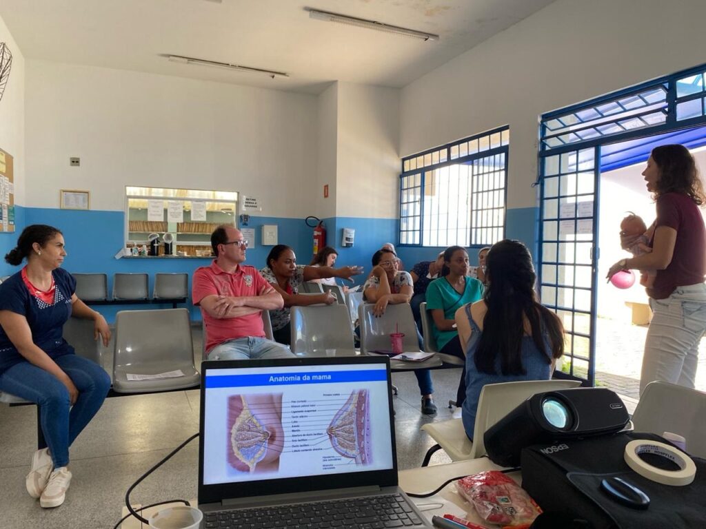 A imagem mostra uma sessão educativa sobre amamentação em uma sala de espera ou clínica de saúde. No centro da cena, uma instrutora, que está de pé e vestindo uma camiseta vinho e calça branca, segura um bebê de brinquedo para demonstrar técnicas de amamentação a um pequeno grupo de adultos sentados em cadeiras de plástico cinza. Entre os participantes, há homens e mulheres, todos atentos à apresentação. Em primeiro plano, há um laptop aberto exibindo uma apresentação em português intitulada "Anatomia da mama", com um diagrama detalhado da estrutura do seio, incluindo termos como pele, músculo peitoral maior, ligamento suspensor, tecido adiposo, lobo, alvéolo, mamilo, ducto lactífero, entre outros. Ao lado do laptop, há um projetor Epson e alguns materiais, possivelmente usados para a palestra. O ambiente é iluminado por luz natural que entra por uma porta aberta e uma janela, e as paredes são pintadas em azul na parte inferior e branco na parte superior. Ao fundo, há um balcão com arquivos e um extintor de incêndio. A atmosfera geral é de aprendizado e cuidado, típica de uma unidade básica de saúde, provavelmente no Brasil, considerando o idioma da apresentação.