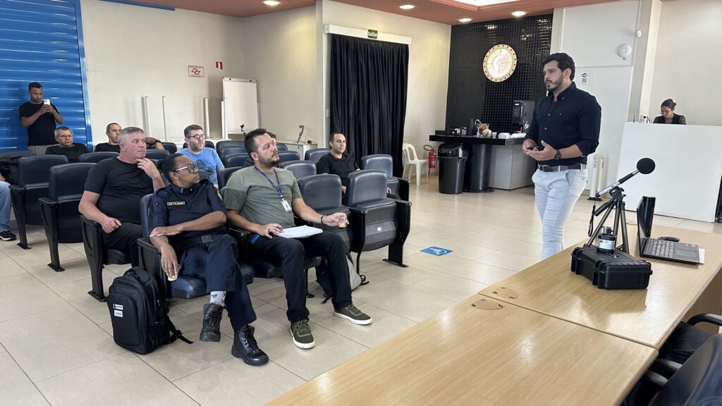 A imagem mostra uma sala de conferências ou auditório com cadeiras azuis, onde um grupo de pessoas está assistindo a uma apresentação. À direita, um homem vestido com uma camisa azul escura e calça clara está em pé, falando e gesticulando, possivelmente o palestrante ou instrutor. Ele está atrás de uma mesa de madeira que tem um notebook, um tripé com microfone e uma caixa preta, que pode conter equipamentos. À esquerda, várias pessoas estão sentadas, prestando atenção ao palestrante. Uma delas usa um uniforme escuro com o nome "GCM ELIANA" bordado, indicando que pode ser uma integrante da Guarda Civil Municipal. Ao fundo, há uma porta com uma cortina preta e uma placa verde escrita "SAÍDA". Também é possível ver uma bancada com uma máquina de café e algumas xícaras, sugerindo uma área de apoio para os participantes. O ambiente é moderno, bem iluminado e organizado, adequado para treinamentos, palestras ou reuniões profissionais.
