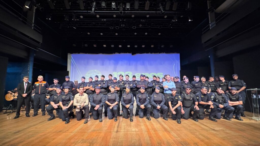 A imagem mostra um grande grupo de pessoas, majoritariamente homens, vestindo uniformes escuros, posando para uma foto em um palco de madeira. Eles estão organizados em várias fileiras, com alguns em pé e outros ajoelhados na frente. A maioria usa uniformes azul escuro ou preto com distintivos, e alguns usam boinas ou bonés. Há também pessoas com coletes táticos. Dois homens à esquerda estão vestidos com roupas civis: um de terno escuro e outro com um colete laranja e preto. Ao fundo, há uma grande tela azul que exibe um logo circular e o texto "100 ANOS", indicando que o evento é uma comemoração de cem anos. O ambiente parece ser uma cerimônia formal, possivelmente relacionada a uma força policial ou grupo de segurança. A iluminação é típica de palco, com refletores visíveis no teto.