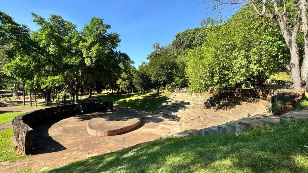 A imagem mostra um anfiteatro ao ar livre, com assentos em degraus feitos de pedra e concreto, formando uma semi-circunferência ao redor de um palco circular. O anfiteatro está situado em um parque verdejante, cercado por muitas árvores altas com folhagem densa. O gramado ao redor é bem cuidado e verde. O céu está claro e azul, indicando um dia ensolarado, provavelmente durante a manhã ou início da tarde. Não há pessoas na cena, o que transmite uma sensação de tranquilidade e paz. Ao fundo, é possível ver mais árvores e alguns elementos de lazer do parque, como mesas de piquenique e brinquedos infantis. O anfiteatro parece ser um local agradável para eventos ao ar livre, cercado pela natureza.