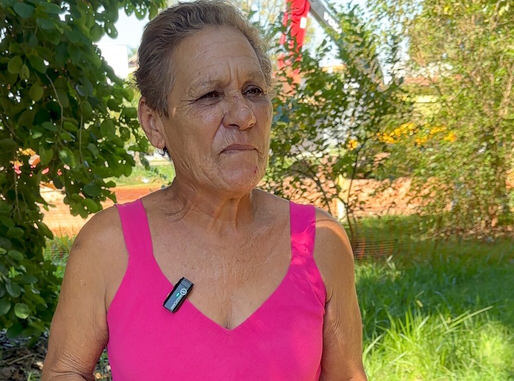 A imagem mostra uma mulher idosa em um plano médio, olhando levemente para o lado direito da câmera. Ela tem cabelos curtos e grisalhos, e sua pele apresenta rugas visíveis, indicando sua idade avançada. Ela veste uma blusa regata rosa vibrante e está usando um microfone de lapela preso na parte da frente da blusa, próximo ao ombro esquerdo. O fundo está desfocado, revelando uma área externa com vegetação verde, possivelmente um parque ou jardim. A luz do sol ilumina suavemente a cena, destacando os detalhes do rosto e da roupa da mulher. A expressão dela é serena, e a composição da foto foca principalmente em sua face e parte superior do corpo, transmitindo uma sensação de tranquilidade em um ambiente natural.