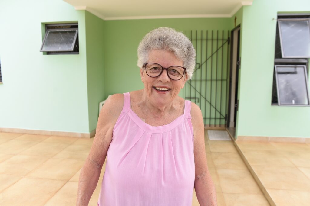 A imagem mostra uma senhora idosa, com cabelos curtos e grisalhos, usando óculos e uma blusa regata rosa. Ela está sorrindo e olhando diretamente para a câmera, transmitindo uma expressão amigável e acolhedora. Ao fundo, vê-se a parede externa de uma casa pintada de verde claro, com duas janelas – uma delas aberta – e uma porta gradeada de metal. O chão é revestido com azulejos claros. A iluminação é natural e intensa, sugerindo que a foto foi tirada durante o dia. A composição foca principalmente na mulher, com o fundo um pouco desfocado, destacando sua figura e expressão.