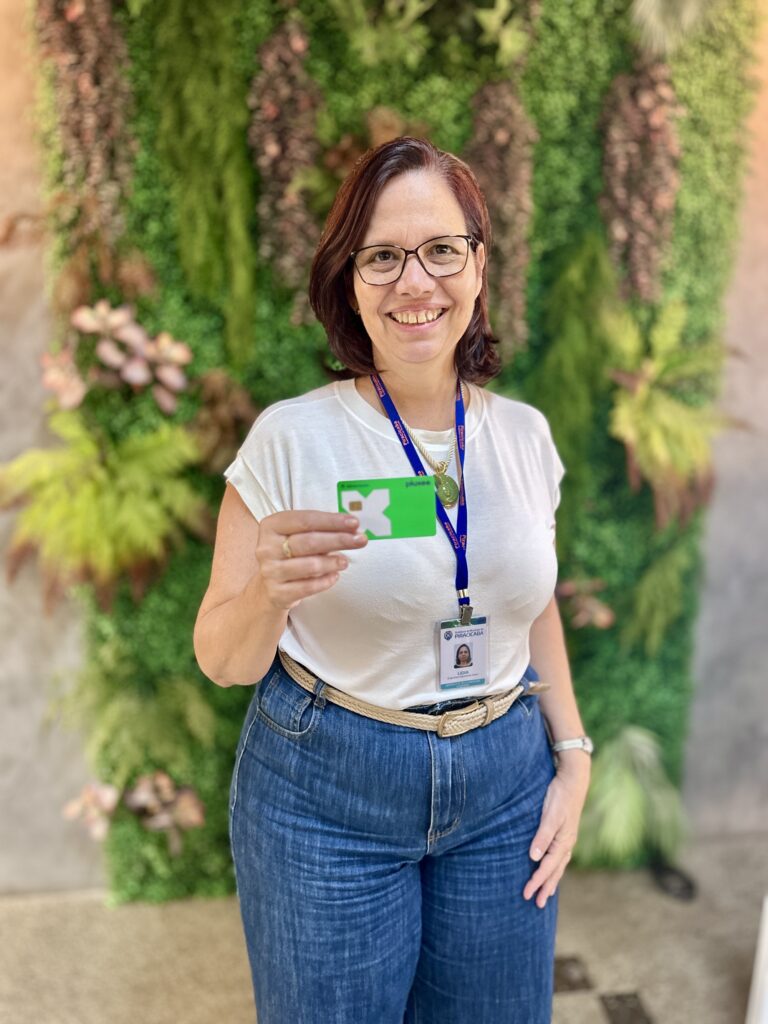 A imagem mostra uma mulher sorridente, em um plano médio, posando em frente a uma parede verde coberta por plantas, que parecem fazer parte de um jardim vertical. Ela está segurando com a mão direita um cartão verde, exibindo-o para a câmera. A mulher usa óculos, uma camiseta branca e calça jeans azul, além de um cinto trançado. Um crachá azul está pendurado no pescoço dela, com identificação que inclui seu nome, "LIDIA", e a profissão "Engenheira Agrônoma", vinculada à "GERENCIA DE PRODUÇÃO E INOVAÇÃO" da "PREFEITURA DO MUNICÍPIO DE PIRACICABA". O ambiente está bem iluminado, com luz natural destacando a pessoa e o cartão, enquanto o fundo de plantas cria um contraste agradável e natural. A imagem transmite uma sensação de profissionalismo e conexão com a natureza.