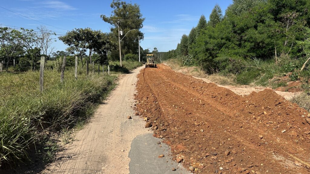 A imagem mostra uma cena rural durante o dia, com céu claro e ensolarado. No centro, há uma máquina amarela, um nivelador de terra (motoniveladora), que está trabalhando na construção ou manutenção de uma estrada de terra vermelha. Ao lado direito da estrada, há um monte de terra recém-movimentada, indicando que o terreno está sendo preparado ou nivelado. À esquerda da estrada, há uma cerca de madeira antiga e postes de energia elétrica com fios que acompanham o caminho. Ao redor da estrada, há vegetação verde, incluindo árvores e arbustos, mostrando que a área é rural e cercada pela natureza. A máquina parece estar se afastando do observador, empurrando a terra para o lado direito da estrada. A imagem transmite uma sensação de trabalho e progresso em um ambiente natural e tranquilo.