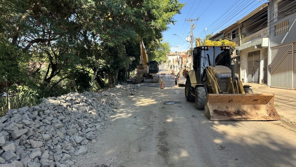 A imagem mostra uma cena urbana onde está ocorrendo uma obra na rua. No centro e à direita, há uma retroescavadeira amarela da marca CAT, destacando-se pela sua grande dimensão e cor vibrante. Mais ao fundo, à esquerda, pode-se ver outra máquina amarela, provavelmente uma escavadeira, que está próxima a uma grande pilha de entulho ou pedras. A rua está parcialmente aberta, com terra e marcas de pneus visíveis, indicando que está em processo de reparo ou pavimentação. Ao redor, há árvores verdes e casas residenciais com muros e portões, sugerindo que a obra está acontecendo em um bairro. O céu está claro e azul, com iluminação natural forte que gera sombras nítidas. A imagem transmite a sensação de trabalho pesado e progresso em infraestrutura local.