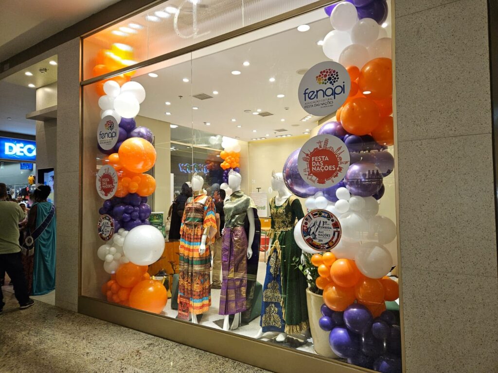 A imagem mostra a vitrine de uma loja decorada para um evento cultural chamado "Festa das Nações". Na vitrine, há manequins vestidos com roupas coloridas e tradicionais, que parecem representar diferentes culturas. Ao redor dos manequins, há muitos balões nas cores laranja, roxo e branco, organizados em grupos e arcos, criando um ambiente festivo e alegre. Também é possível ver vários cartazes circulares com textos relacionados ao evento, como "Festa das Nações", "Associação Cultural", "Fenapi" e "Corrida das Nações". O ambiente parece ser um shopping, já que há outras lojas visíveis ao fundo, incluindo uma com a placa "DEC". A iluminação é clara e destaca bem a vitrine e a decoração. No geral, a imagem transmite uma atmosfera de celebração multicultural, com foco em roupas típicas, elementos decorativos vibrantes e informações sobre atividades relacionadas à festa.