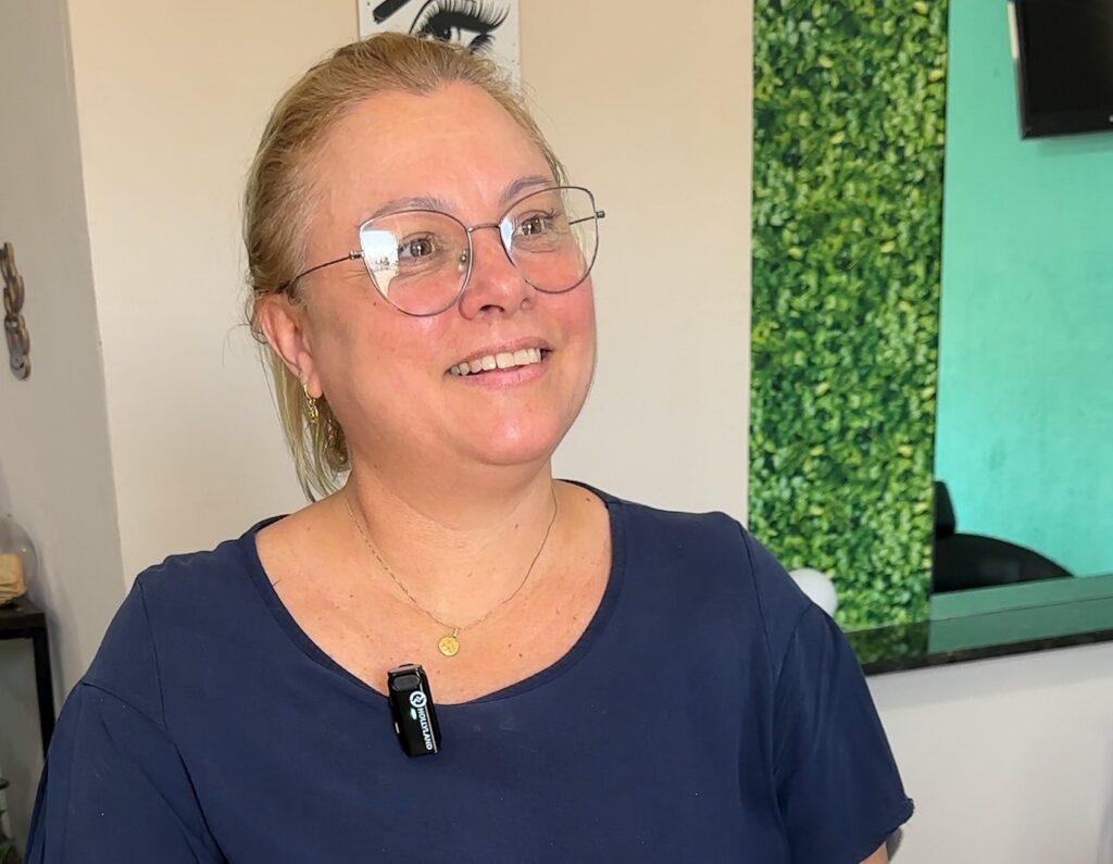 A imagem mostra uma mulher de meia-idade, com cabelos loiros presos para trás, usando óculos e uma camiseta azul escura. Ela está sorrindo levemente e olhando para o lado, em direção à direita da imagem. Ela usa um colar com um pingente dourado e um microfone de lapela preso à camiseta. Ao fundo, há uma parede com uma seção vertical de folhagem verde artificial e, ao lado, uma parede azul turquesa com um objeto preto que parece ser uma tela ou monitor. A iluminação é clara e uniforme, destacando o rosto da mulher. A imagem transmite uma sensação de descontração e simpatia.