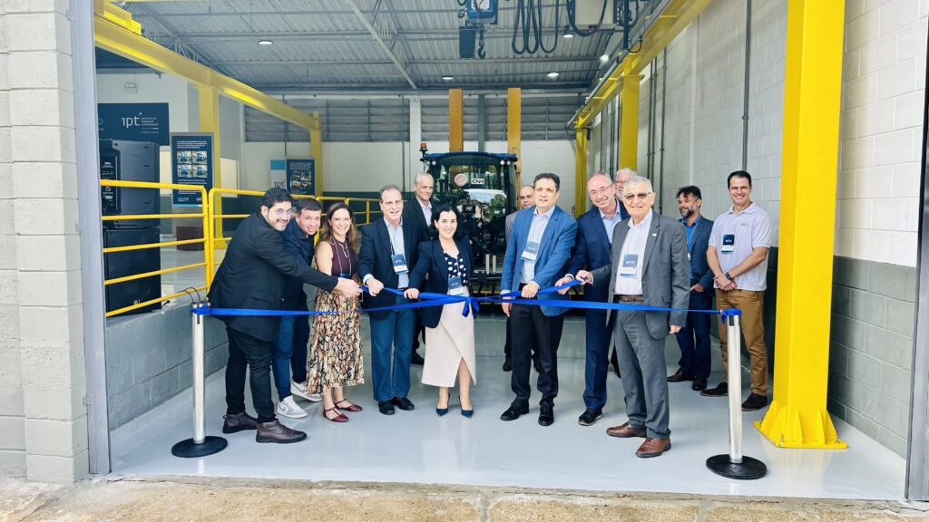 A imagem mostra uma cerimônia de inauguração ou lançamento dentro de uma instalação industrial, provavelmente uma fábrica ou centro de pesquisas, dado o ambiente e os equipamentos ao fundo. No centro da imagem, há um grupo de doze pessoas, entre homens e mulheres, todas vestidas de forma formal ou casual executiva. Uma mulher no centro segura uma fita azul, indicando que está prestes a ser cortada, simbolizando a abertura oficial do evento ou espaço. Ao fundo, destaca-se um trator azul grande, identificado com a marca "CNH", que é um fabricante conhecido de equipamentos agrícolas e de construção. O local tem paredes de concreto e estruturas metálicas amarelas, típicas de ambientes industriais. Também são visíveis placas com o logo do "ipt" (Instituto de Pesquisas Tecnológicas), sugerindo que o evento está relacionado a essa instituição, possivelmente uma parceria ou lançamento de um projeto tecnológico ligado ao setor agrícola ou industrial.