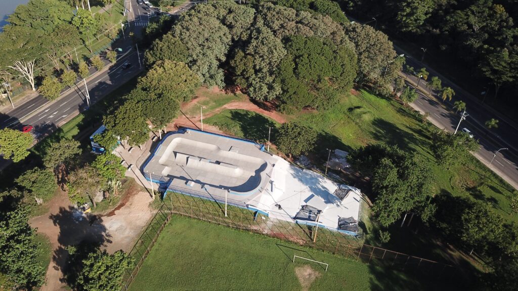 A imagem mostra uma vista aérea de um skatepark moderno, situado em um parque cercado por muitas árvores verdes e um gramado bem cuidado. O skatepark é feito de concreto e possui várias rampas, bowls e obstáculos para a prática de skate. Algumas áreas estão cobertas por lonas pretas, indicando obras ou manutenção. Várias pessoas estão no local, algumas andando de skate e outras observando. Ao redor do parque, há uma cerca de arame que separa o skatepark de um campo de futebol com um gol visível. Próximo ao parque, há uma estrada com alguns carros e postes de luz. O dia está ensolarado, o que cria sombras nítidas no chão, e a vegetação ao redor é densa e verde. A imagem é clara e colorida, mostrando detalhes da estrutura do skatepark e do ambiente natural e urbano ao redor.