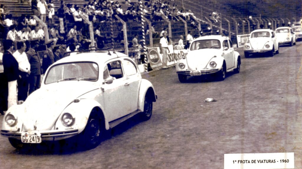 A imagem mostra uma cena histórica, provavelmente de 1960, conforme indicado pelo texto "1ª FROTA DE VIATURAS - 1960" no canto inferior direito. Nela, uma fila de carros Volkswagen Fusca brancos está alinhada em uma pista ou estrada, possivelmente durante um evento oficial ou desfile. Os carros parecem estar em ótimo estado, sugerindo que fazem parte de uma frota organizada, talvez de veículos oficiais ou de patrulha. Ao fundo, há arquibancadas cheias de pessoas assistindo ao evento, o que reforça a ideia de uma ocasião pública e importante. A presença de placas com a palavra "BRAHMA" indica que pode haver patrocínio ou alguma relação com a marca, comum em eventos esportivos ou sociais da época. A fotografia tem um tom envelhecido, típico de imagens antigas, com cores amareladas e um aspecto granuloso, o que contribui para a sensação de nostalgia. A composição da foto, com os carros em perspectiva e as arquibancadas ao fundo, transmite a organização e a solenidade do momento registrado.
