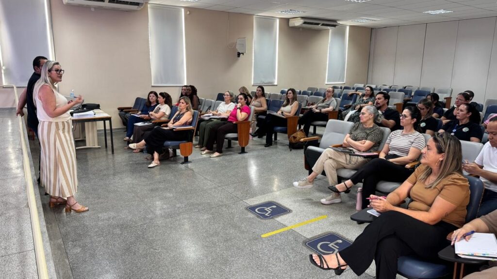A imagem mostra uma apresentação ou palestra ocorrendo em um auditório ou sala de aula de tamanho médio. Uma mulher loira, vestindo uma blusa branca sem mangas e calça listrada larga, está em pé na frente, à esquerda, gesticulando enquanto fala para a audiência. Atrás dela, há um homem de camiseta preta parcialmente visível. A audiência é composta por cerca de 20 a 25 adultos, principalmente mulheres, sentados em cadeiras estofadas azuis e cinzas organizadas em fileiras. Alguns participantes estão anotando, enquanto outros escutam atentamente. O ambiente tem paredes de cor neutra, piso cinza com manchas, e iluminação fluorescente no teto. Há várias janelas grandes com cortinas nas paredes laterais e ao fundo, além de aparelhos de ar-condicionado instalados próximo ao teto. No chão, em primeiro plano, há dois símbolos azuis indicando acessibilidade para cadeirantes, separados por uma linha amarela. A foto foi tirada em ângulo amplo, levemente elevado e lateral, capturando tanto a palestrante quanto a maioria da audiência. A iluminação é clara e uniforme, típica de ambientes internos profissionais ou educacionais. A qualidade técnica da imagem é boa, com foco nítido e profundidade de campo que mantém todos os elementos visíveis.