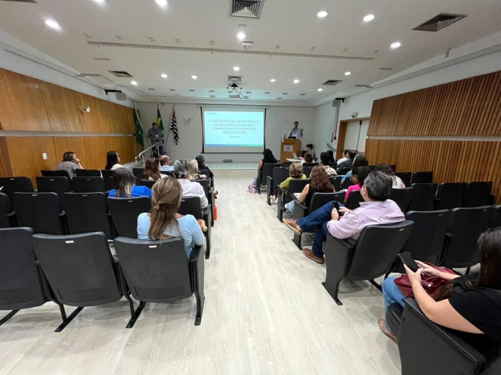 A imagem mostra uma sala de conferências ou auditório onde está acontecendo uma apresentação. Na frente da sala, há uma tela de projeção exibindo um slide com o título "PLANO MUNICIPAL DE PROMOÇÃO DA CONVIVÊNCIA FAMILIAR E COMUNITÁRIA DE CRIANÇAS E ADOLESCENTES 2024-2938". Dois apresentadores estão na frente, um próximo à tela e outro em um púlpito. O público, composto por cerca de 30 a 40 pessoas, está sentado em cadeiras organizadas em fileiras, a maioria são mulheres. A sala tem paredes com painéis de madeira clara e iluminação artificial proveniente de luminárias no teto. A atmosfera parece formal ou semi-formal, típica de uma reunião, seminário ou sessão educativa. A imagem foi capturada de um ângulo ligeiramente elevado, mostrando uma boa parte do espaço e dos participantes.