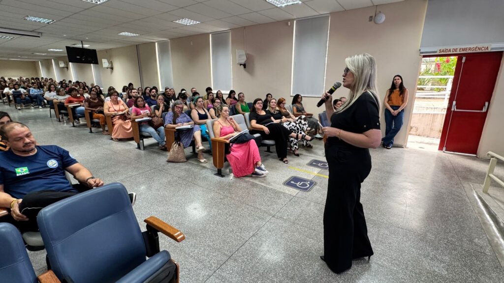 A imagem mostra uma mulher loira, vestindo uma roupa preta, falando ao microfone em um auditório. Ela está de pé à direita da imagem, aparentemente fazendo uma apresentação ou palestra para um público. O auditório está cheio de pessoas sentadas em cadeiras estilo teatro, a maioria delas mulheres, que parecem estar prestando atenção à fala. No canto esquerdo da imagem, um homem usa uma camiseta azul com a palavra "BRASIL" escrita nas costas. Ao fundo, há uma porta vermelha com uma placa acima indicando "SAÍDA DE EMERGÊNCIA". A iluminação é clara, típica de ambientes internos. A cena transmite um ambiente formal e organizado, possivelmente um evento educacional, profissional ou cultural.