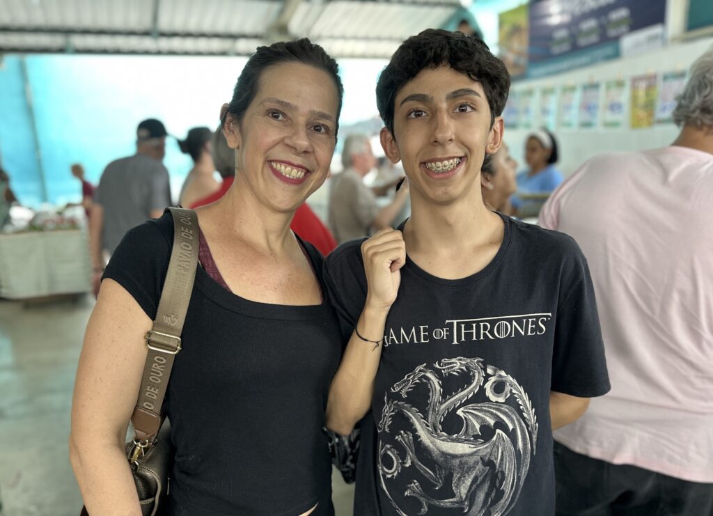 A imagem mostra uma mulher e um adolescente sorrindo, posando para uma foto em um ambiente ao ar livre que parece ser uma feira ou mercado coberto. Ambos estão vestidos de forma casual: a mulher usa uma blusa preta de manga curta e carrega uma bolsa marrom com uma alça que tem a inscrição "PINHÃO DE OURO". O adolescente veste uma camiseta preta com a estampa da série "Game of Thrones" e usa aparelho nos dentes. Ao fundo, é possível ver outras pessoas e barracas, com uma cobertura metálica e lonas azuis, indicando um local de comércio informal ou um evento comunitário. A expressão dos dois é alegre e descontraída, transmitindo um momento de convivência e diversão.