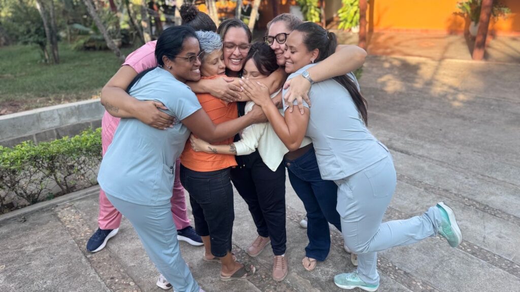 A imagem mostra um grupo de sete mulheres abraçadas em um círculo apertado, demonstrando afeto e amizade. Elas estão ao ar livre, em um espaço que parece ser um jardim ou pátio, com árvores e plantas ao fundo, além de uma parede laranja de um prédio. O chão é de pedra e concreto. As mulheres têm idades e etnias variadas, e estão sorrindo, transmitindo felicidade e união. Algumas vestem uniformes, possivelmente de trabalho, em tons de azul claro e rosa, enquanto outras estão com roupas casuais, como camisetas e jeans. A luz do dia ilumina suavemente a cena, sugerindo que é fim de tarde. A imagem transmite uma sensação de calor humano, companheirismo e alegria.