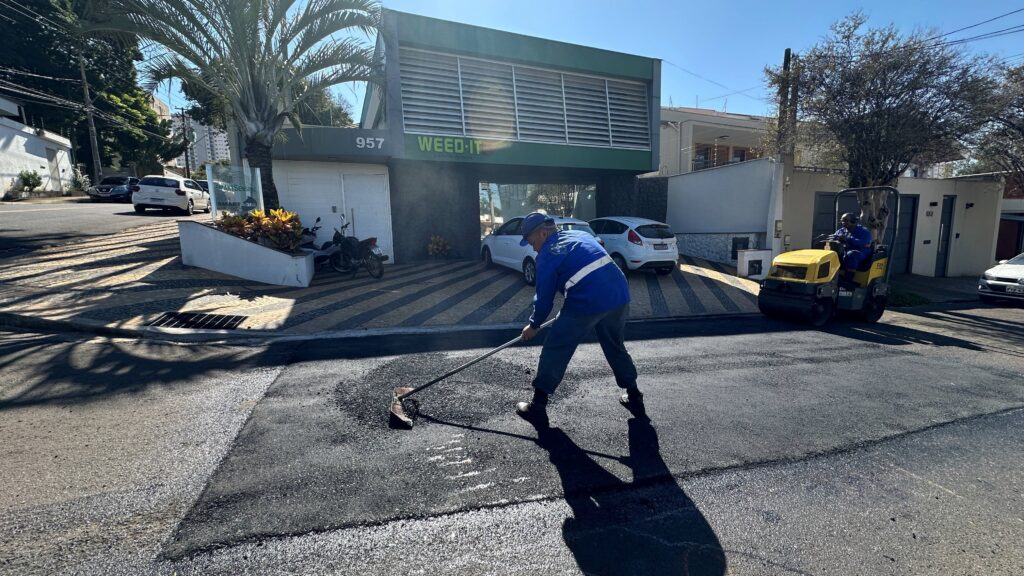 A imagem mostra uma cena de reparo de estrada em um dia ensolarado. Em primeiro plano, há um trabalhador vestido com uniforme azul e faixas refletivas, usando um ancinho para espalhar asfalto fresco. Atrás dele, uma máquina compactadora amarela está sendo operada por outro trabalhador também de uniforme azul, indicando um trabalho coordenado na pavimentação. A superfície da estrada apresenta uma mistura de asfalto antigo, asfalto novo e cascalho. Ao fundo, há um edifício moderno com o número "957" e a inscrição "WEED-IT" em destaque, além dos textos "SMART SENSING" e "SMART AGRI". Alguns carros estão estacionados ou trafegando pela rua, e uma motocicleta está próxima à entrada do edifício. A área ao redor parece ser residencial ou comercial, com casas e postes de luz visíveis. A luz do sol forte cria sombras definidas de palmeiras e postes, gerando um padrão de luz e sombra no chão e na calçada. A imagem transmite uma sensação de atividade urbana e trabalho em andamento.