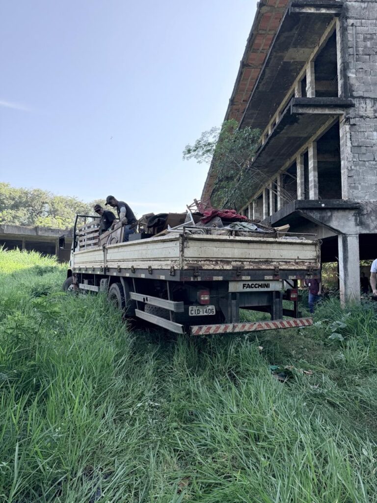 A imagem mostra um caminhão plataforma carregado com entulhos, estacionado em um terreno coberto por grama alta e verde. No compartimento de carga do caminhão, há duas pessoas, aparentemente trabalhando na organização ou remoção dos materiais. Ao fundo, destaca-se um prédio inacabado ou abandonado, construído em concreto e tijolos aparentes, com várias lajes expostas e estruturas metálicas visíveis. A vegetação cresce ao redor e até sobre partes do prédio, indicando que ele está sem manutenção há algum tempo. O caminhão é branco e tem a marca "FACCHINI" visível na parte traseira, com a placa de identificação "CID-1406". O cenário sugere um local rural ou uma área que está passando por limpeza ou recuperação, dado o estado do prédio e a presença de entulho. A luz natural do dia ilumina a cena, com céu claro e sombras definidas. A composição da imagem chama atenção para o contraste entre a natureza que cresce livremente e a estrutura humana abandonada.