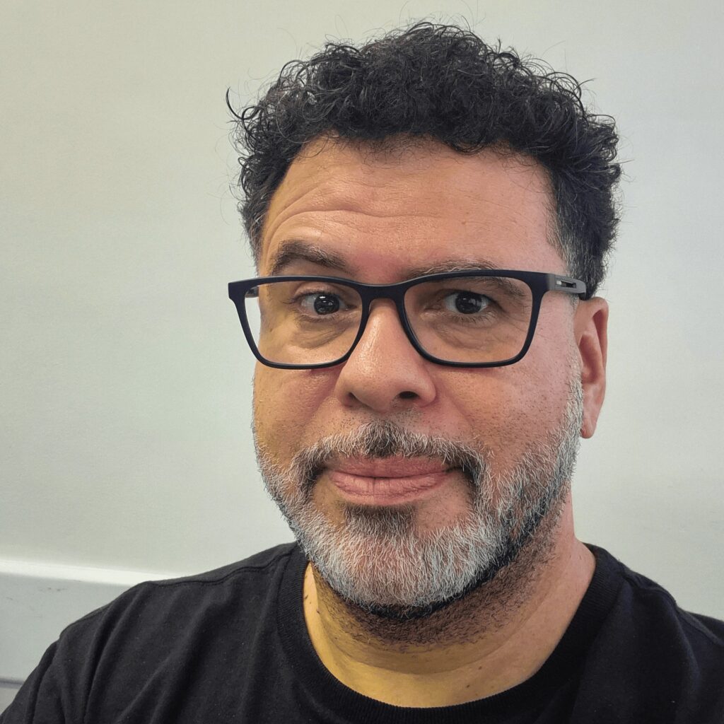 A imagem mostra um homem de meia-idade, com cabelos escuros e cacheados, além de uma barba grisalha. Ele usa óculos de aro preto e uma camiseta preta. O fundo é claro e neutro, provavelmente uma parede interna, e a iluminação é uniforme, destacando bem seu rosto. A expressão dele é calma, com um leve sorriso. A foto é uma selfie ou retrato próximo, focando principalmente no rosto do homem.