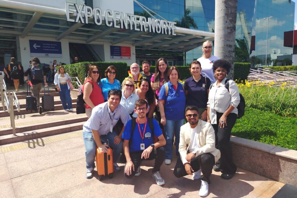 A imagem mostra um grupo de cerca de 15 pessoas reunidas em frente à entrada do prédio chamado "EXPO CENTER NORTE". O grupo está posicionado em duas fileiras, com algumas pessoas em pé e outras agachadas na frente, todas sorrindo e aparentando estar felizes. O local parece ser um centro de convenções ou eventos, com uma fachada moderna de vidro refletindo a luz do sol. O ambiente está iluminado pela luz natural do dia, com sombras evidentes no chão, indicando um céu claro. Em primeiro plano, há um piso pavimentado e algumas plantas com flores amarelas. Algumas pessoas carregam malas e mochilas, sugerindo que estão participando de um evento ou viagem. Acima da entrada, há placas indicando diferentes pavilhões ("Pavilhão Branco", "Pavilhão Azul") e a área de credenciamento. A atmosfera geral é de descontração e confraternização.