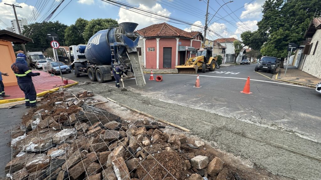 A imagem mostra uma cena de construção em uma rua residencial durante o dia, com céu parcialmente nublado. O foco principal é o despejo de concreto úmido de um caminhão betoneira azul sobre uma área em construção na rua. Vários trabalhadores com uniformes azuis escuros e faixas amarelas de alta visibilidade estão envolvidos na obra; um deles está utilizando uma pá para espalhar o concreto, enquanto outro observa o trabalho. O concreto fresco forma uma camada cinza sobre a rua, cercada por uma área com pedras, terra e uma malha metálica. Ao fundo, há uma retroescavadeira amarela estacionada, além de alguns carros e caminhões. As casas ao redor apresentam telhados vermelhos e paredes pintadas em tons avermelhados. Cone de sinalização laranja delimita a área da obra. A iluminação indica um dia ensolarado, com sombras evidentes, e a perspectiva da foto é ligeiramente inclinada para cima, destacando o caminhão e o céu. A imagem transmite a sensação de um trabalho de infraestrutura em andamento em um bairro residencial.