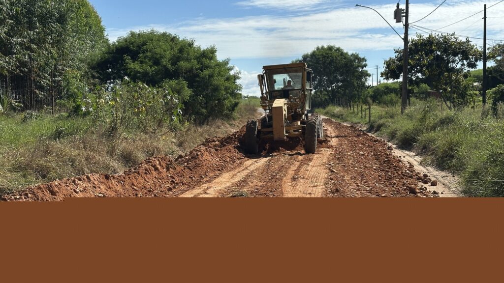 A imagem mostra uma motoniveladora amarela no centro, trabalhando na construção de uma estrada de terra. A máquina está em uma estrada de terra vermelha, nivelando o solo. À esquerda, há vegetação verde densa, com árvores e arbustos, enquanto à direita há uma cerca e postes de energia elétrica com cabos. O céu está claro, azul com algumas nuvens finas, indicando um dia ensolarado. A foto é tirada de um ângulo baixo, o que destaca o tamanho da motoniveladora. O operador está visível dentro da cabine da máquina, que é fechada por vidros. A cena transmite uma sensação de trabalho em andamento em uma área rural ou semi-rural.