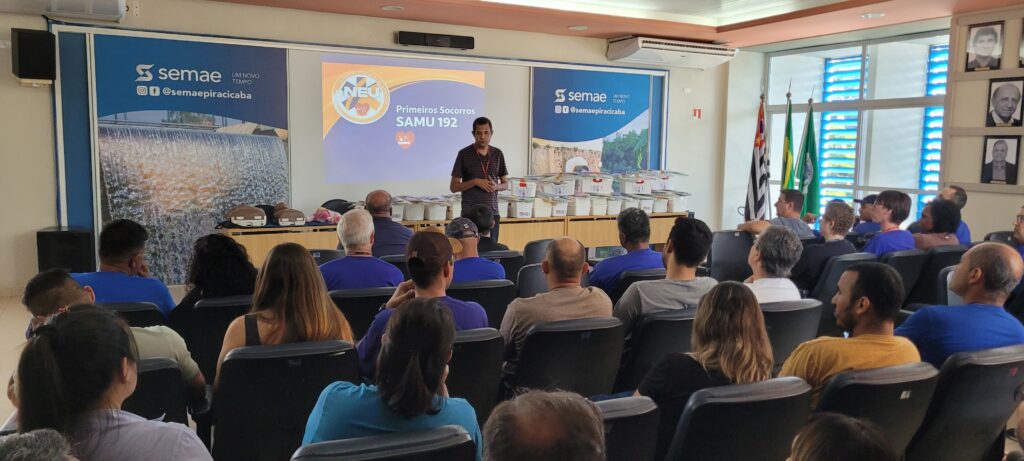 A imagem mostra uma sala de treinamento ou palestra, onde um homem está em pé na frente, falando para um grupo de pessoas sentadas em cadeiras. Ele parece ser o instrutor ou palestrante e está gesticulando enquanto fala. Atrás dele, há uma grande tela com uma apresentação exibida, contendo textos e logos relacionados a "Primeiros Socorros" e "SAMU 192". Nas laterais da tela, há banners grandes com a marca "semae" e imagens relacionadas a água e infraestrutura. À direita, há várias bandeiras, incluindo a do Brasil e do estado de São Paulo, além de quadros na parede. Também é possível ver caixas brancas empilhadas em uma mesa, possivelmente materiais para o treinamento. O público está atento ao palestrante, e a sala é bem iluminada, com ar-condicionado no teto. A atmosfera indica um ambiente formal e educacional, voltado para capacitação em primeiros socorros, provavelmente organizado pela semae de Piracicaba.