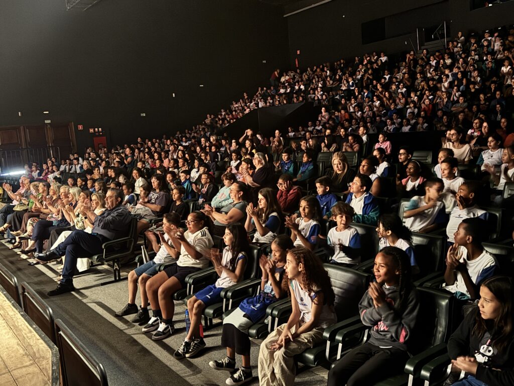 A imagem mostra um grande auditório ou teatro cheio de pessoas sentadas, principalmente crianças e jovens, com alguns adultos. Elas estão organizadas em fileiras de cadeiras escuras, olhando para o palco (que não aparece na foto). Muitas pessoas estão aplaudindo, demonstrando que estão apreciando uma apresentação ou espetáculo. A iluminação está baixa, com foco na plateia, criando um ambiente acolhedor e envolvente. No fundo, há placas de saída de emergência com a palavra "SAÍDA", indicando que o local segue normas de segurança. A cena transmite uma sensação de união e entusiasmo dos espectadores diante do evento.