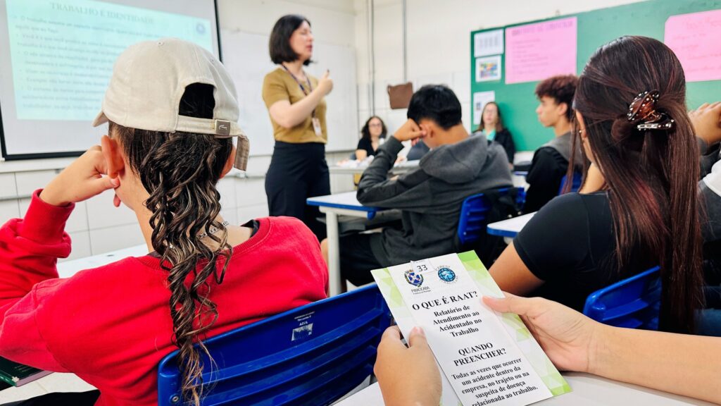 A imagem mostra uma sala de aula com várias pessoas sentadas, aparentemente participando de uma palestra ou workshop. Em primeiro plano, há uma pessoa de costas, usando um boné branco e um moletom vermelho. Essa pessoa está segurando um folheto intitulado "O QUE É RAAT?", que explica o Relatório de Atendimento ao Acidentado no Trabalho, destacando quando deve ser preenchido (sempre que ocorrer um acidente na empresa, no trajeto ou suspeita de doença relacionada ao trabalho). Ao fundo, uma mulher está em pé, de frente para os alunos, falando e apontando para um projetor que exibe um slide com o título "TRABALHO E IDENTIDADE". O texto do slide discute como o trabalho influencia a identidade da pessoa, mencionando que o trabalho é o que a pessoa faz e produz, e que a falta de resultados pode gerar sofrimento e isolamento. A sala tem paredes brancas, um quadro verde com avisos colados, e cadeiras azuis. A atmosfera é de aprendizado e reflexão sobre a relação entre trabalho e identidade, além de questões de saúde e segurança no trabalho.