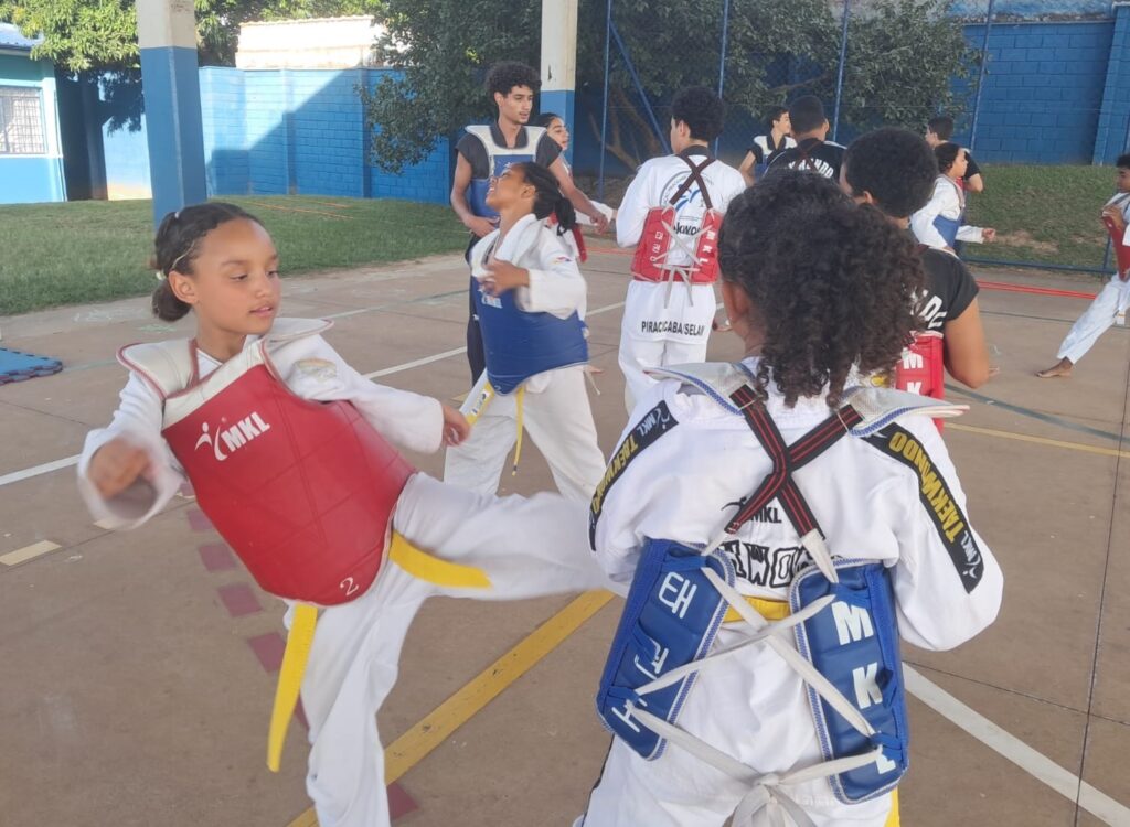 A imagem mostra um grupo de crianças e adolescentes praticando Taekwondo em um espaço aberto, possivelmente um pátio escolar ou centro comunitário. O chão é de concreto com marcações de quadra esportiva. No centro da imagem, uma menina está executando um chute alto, vestindo um uniforme branco de Taekwondo (dobok), um protetor peitoral vermelho com a inscrição "MKL" e uma faixa amarela na cintura. À sua frente, outra menina com o uniforme branco e um protetor azul, com a palavra "TAEKWONDO" escrita nas mangas, parece estar pronta para receber ou bloquear o chute. Ao fundo, há outros praticantes, alguns com protetores vermelhos e outros com azuis, além de um adulto ou monitor vestido com camiseta preta. As paredes ao redor são pintadas de azul, e há árvores e gramado ao fundo, indicando que o local é ao ar livre. Na parte inferior do protetor de algumas crianças, está escrito "PIRACICABA/SELAN", sugerindo que a atividade acontece em Piracicaba, Brasil. A imagem transmite uma sensação de concentração, disciplina e energia típica de um treino de artes marciais.