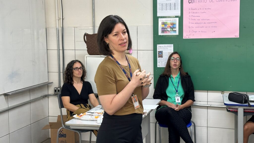 A imagem mostra uma sala de aula ou ambiente escolar, onde três mulheres estão presentes. A mulher no centro está em pé, parece estar falando ou apresentando algo, vestindo uma camiseta amarela mostarda e calça preta, com um crachá pendurado no pescoço. As outras duas mulheres estão sentadas, uma com cabelo cacheado e óculos, vestindo uma blusa preta, e a outra com cabelo liso, também de óculos, usando uma blusa verde com um casaco preto. Ao fundo, há um quadro verde com um cartaz rosa intitulado "CONTRATO DE CONVIVÊNCIA", que lista regras como respeito, empatia, maturidade, autorização para saída, uso pedagógico dos equipamentos, compromisso com os estudos e pontualidade. Também há recortes de jornal escolar e uma tabela colorida, provavelmente um cronograma ou horário. O ambiente é iluminado por luz artificial, com paredes brancas e azulejos, e um quadro branco à esquerda. A atmosfera parece ser de uma reunião ou apresentação escolar, com foco em educação e convivência.