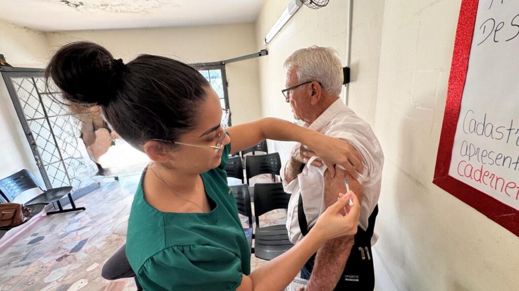 A imagem mostra uma cena de vacinação em um ambiente interno, possivelmente uma clínica ou centro comunitário. Um profissional de saúde, vestindo um uniforme verde, está aplicando uma vacina no braço direito de um homem idoso, que usa uma camisa branca de manga curta e óculos. O idoso está sentado em uma cadeira preta, enquanto a profissional está em pé ao seu lado, concentrada no procedimento. O ambiente é iluminado por luz natural que entra pela esquerda. Ao fundo, há outras cadeiras pretas e uma parede clara com um quadro branco preso, onde se lê parcialmente as palavras "De S", "Cadastr", "apresent" e "Cadernet", sugerindo termos relacionados a cadastro e caderneta de vacinação. O homem idoso tem uma bolsa preta com a palavra "SPORTS" escrita. A cena transmite um momento de cuidado e prevenção, típico de campanhas de vacinação, com atenção e respeito ao paciente.